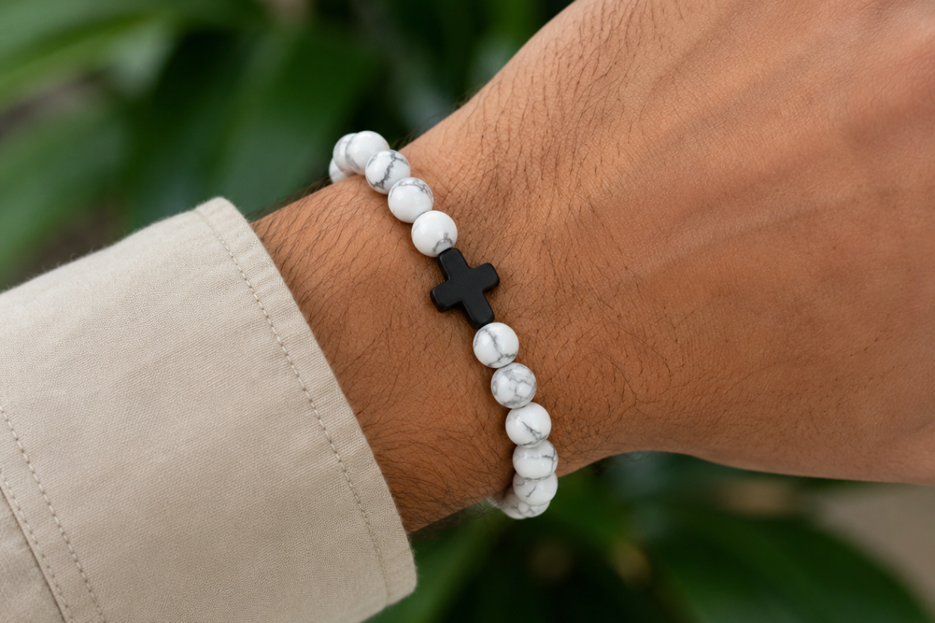 Bracelet en Howlite blanche avec croix noire porté au poignet, avec perles blanches veinées devant un décor végétal.