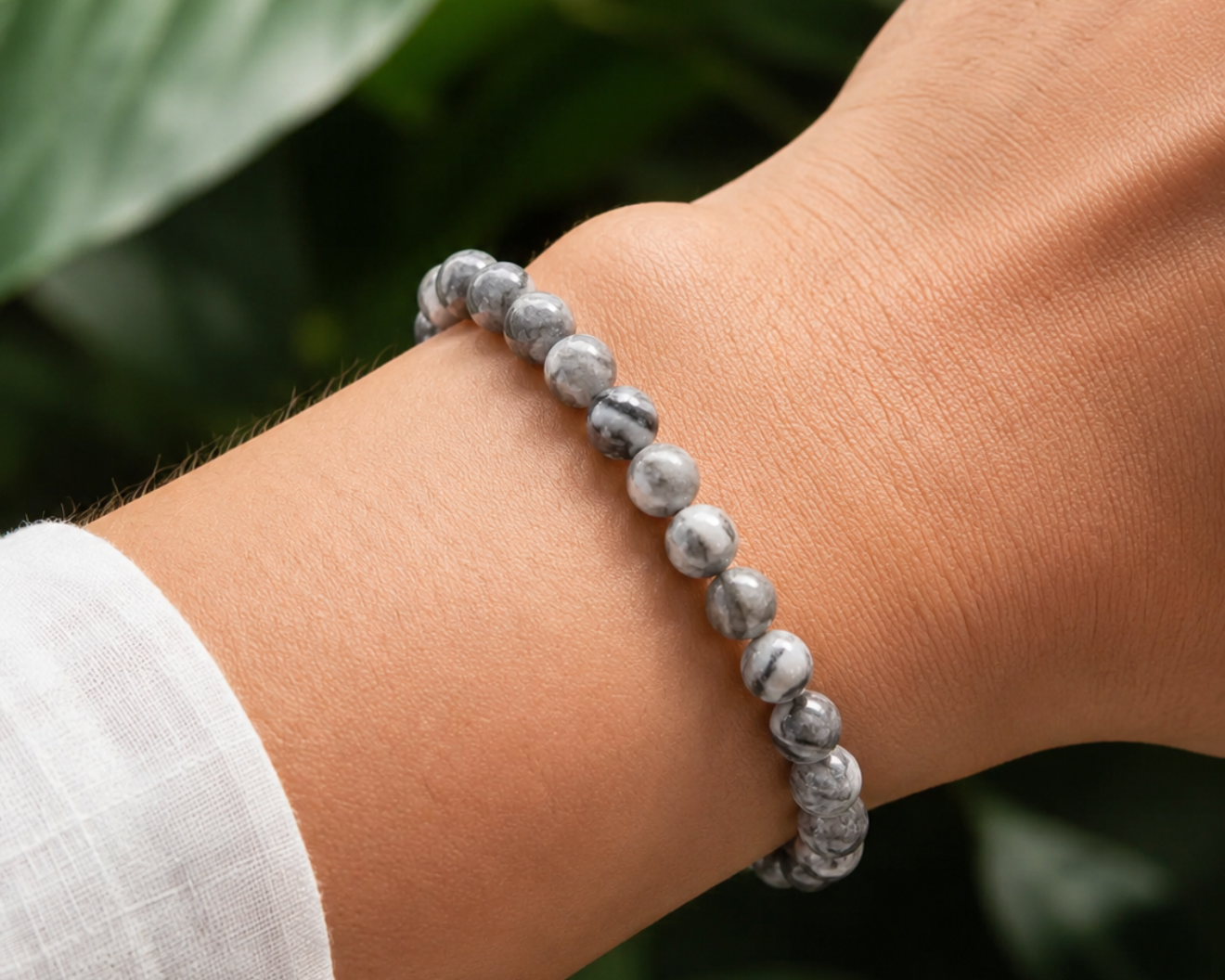 Bracelet en Jaspe Picasso porté au poignet, avec perles grises en pierre naturelle aux motifs marbrés devant un décor végétal.
