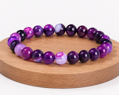 Bracelet en Agate rubanée traitée violette présenté sur un socle en bois, avec perles violettes marbrées sur fond blanc.