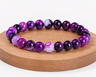 Bracelet en Agate rubanée traitée violette présenté sur un socle en bois, avec perles violettes marbrées sur fond blanc.