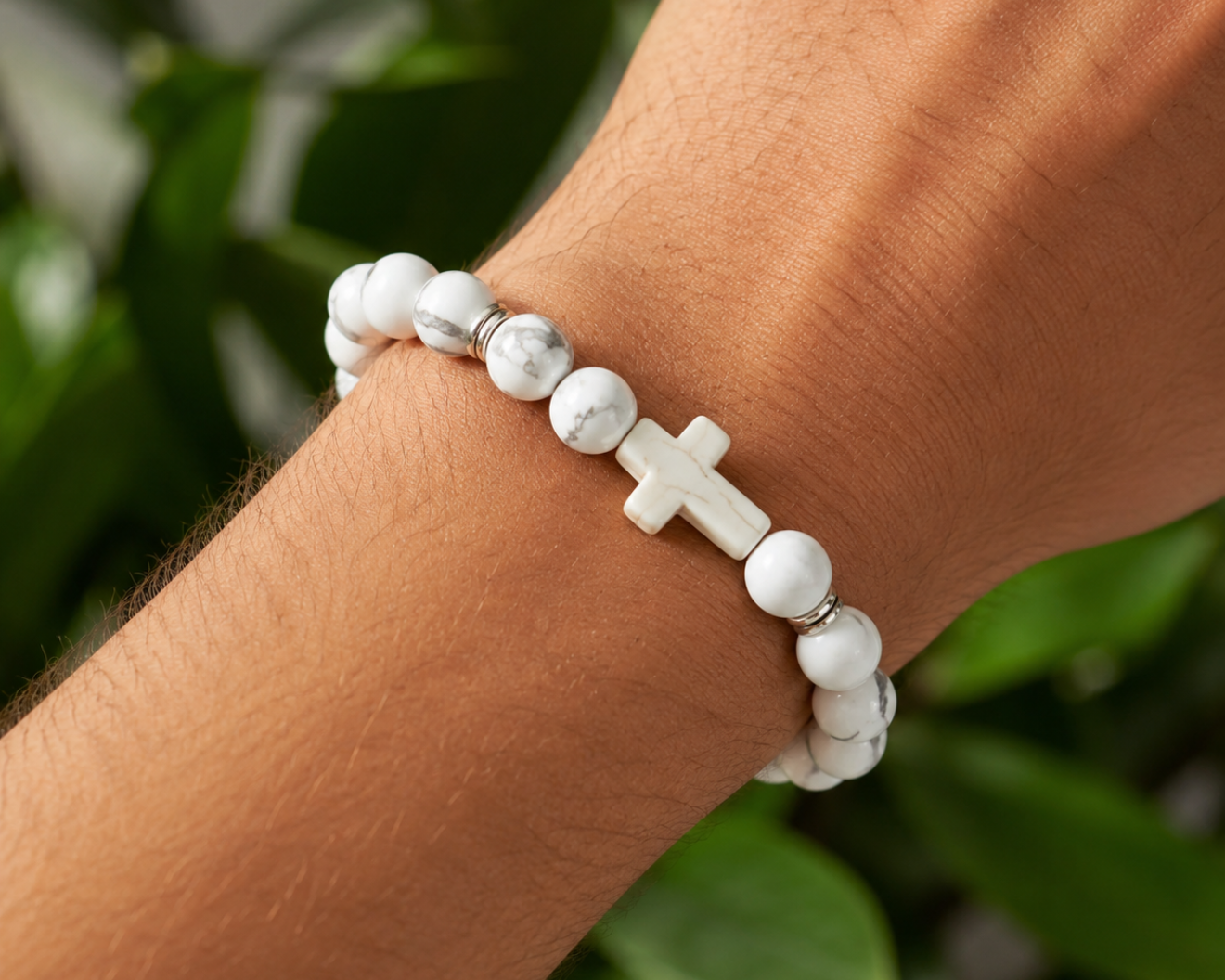 Bracelet en Howlite blanche avec croix blanche porté au poignet, avec perles blanches veinées et détails métal devant un décor végétal.