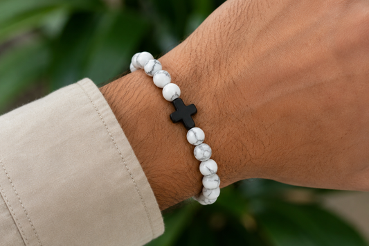 Bracelet en Howlite blanche avec croix noire porté au poignet, avec perles blanches veinées devant un décor végétal.