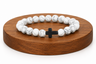 Bracelet en Howlite blanche avec croix noire présenté sur un socle en bois fond blanc, avec perles blanches veinées.