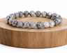 Bracelet en Jaspe Picasso présenté sur un socle en bois fond blanc, avec perles grises en pierre naturelle aux motifs marbrés.