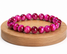 Bracelet en Œil de Tigre traité fuchsia présenté sur un socle en bois, avec perles chatoyantes sur fond blanc.