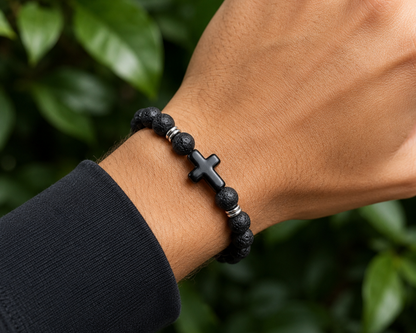 Bracelet en Pierre de Lave avec croix noire et perles métal porté au poignet, avec perles noires texturées devant un décor végétal.