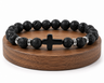 Bracelet en Pierre de Lave avec croix noire et perles métal, présenté sur un socle en bois fond blanc avec perles noires texturées.