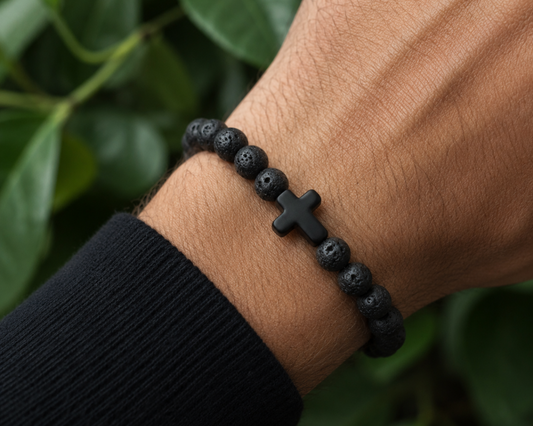 Bracelet en Pierre de Lave avec croix noire porté au poignet, avec perles noires texturées devant un décor végétal.