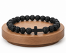 Bracelet en Pierre de Lave avec croix noire présenté sur un socle en bois fond blanc, avec perles noires texturées.